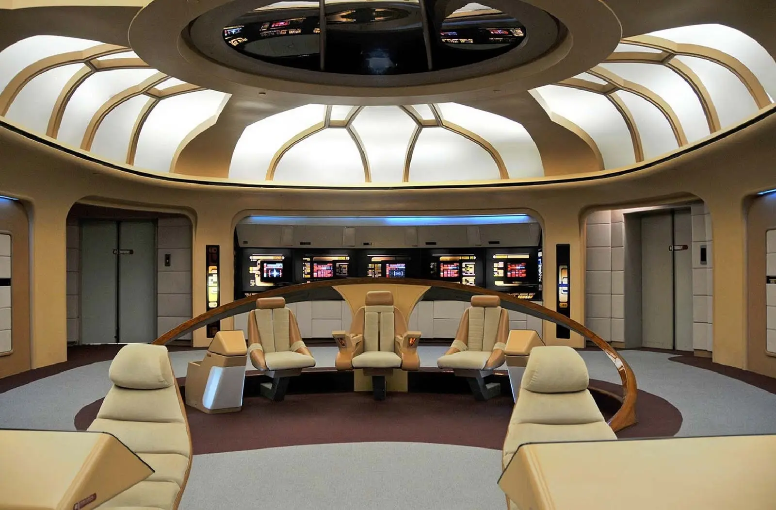 The bridge of a futuristic starship, designed with a circular seating arrangement. In the center, a large, command chair is surrounded by four other seats, all with ergonomic, cream-colored upholstery. The curved ceiling has a series of geometric panels with ambient lighting. Behind the seats, large control consoles with touch-screen interfaces are mounted on the walls. Two doorways and an elevator are visible on either side of the bridge.