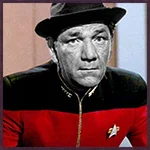 Black-and-white image of a man wearing a red and black Starfleet uniform with a black hat. He has a serious expression and is looking forward, with a neutral background