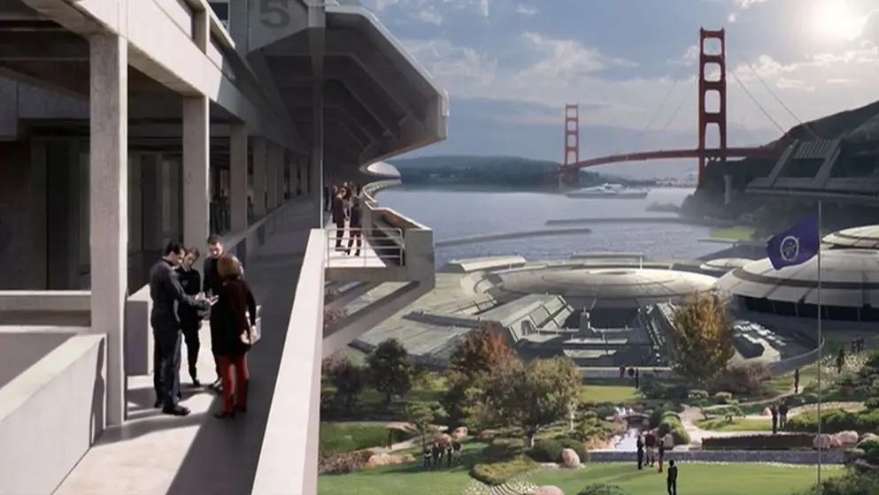 A futuristic campus scene featuring people conversing on a balcony overlooking a lush courtyard with trees, pathways, and landscaped gardens. In the distance, domed buildings and a flag bearing the emblem of the United Federation of Planets are visible near the waterfront. The Golden Gate Bridge spans the background under a partly cloudy sky.