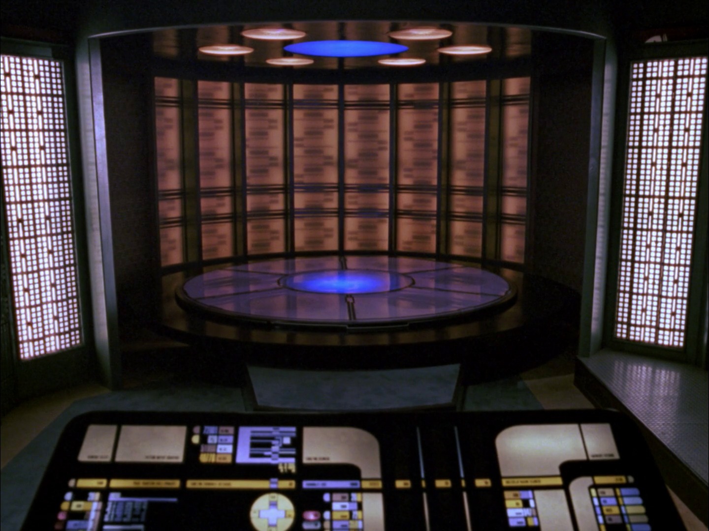 A futuristic transporter room from a starship, featuring a circular transport pad with glowing blue lights and a surrounding control console displaying LCARS interface. The walls are lined with illuminated panels, and overhead lights highlight the center of the pad.