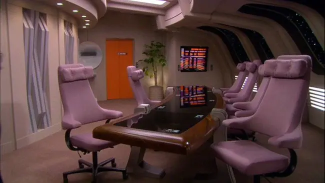 A futuristic starship conference room with high-backed, lavender chairs surrounding a sleek black and wood table. The room features large windows revealing stars outside, wall-mounted display panels with LCARS interface, and a potted plant near a bright orange door.