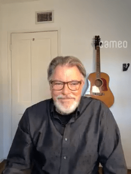 A man with gray hair and beard, wearing glasses and a dark button-up shirt, sits in a well-lit room with a wooden acoustic guitar hanging on the wall behind him. This is Actor Jonathan Frakes.