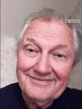 Actor Andrew Robinson smiling warmly in a close-up selfie video. He is wearing a dark shirt and is positioned in front of a softly lit, neutral-toned wall with a subtle pattern. The word 'Cameo' appears in the top right corner.