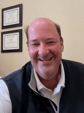 Actor Brian Baumgartner smiling in a well-lit office setting. He is wearing a white polo shirt under a dark vest, with framed certificates or diplomas hanging on the wall behind him.