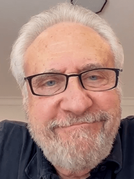 Actor Brent Spiner smiling subtly while wearing glasses and a dark shirt, with a neatly trimmed white beard and light gray hair, seated indoors in a softly lit room.
