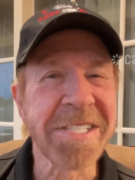 Actor and martial artist Chuck Norris smiling while wearing a black cap and dark polo shirt, seated indoors in front of windowed doors with warm lighting.