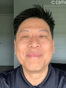 Actor Garrett Wang smiling gently while wearing a dark collared shirt, standing in a brightly lit room with white walls and a closed door behind him.