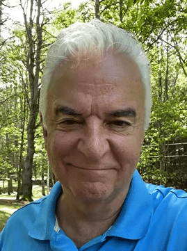 Actor John O'Hurley smiling outdoors in a wooded area, wearing a light blue polo shirt with sunlight filtering through the trees behind him.