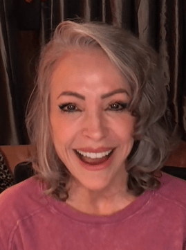 Actress Nana Visitor smiling warmly, wearing a pink top with styled gray hair, seated indoors in front of dark curtains.