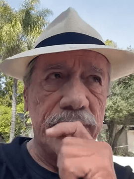 Actor Edward James Olmos outdoors wearing a white fedora with a dark band, thoughtfully resting his hand near his mouth, with palm trees and greenery in the background.