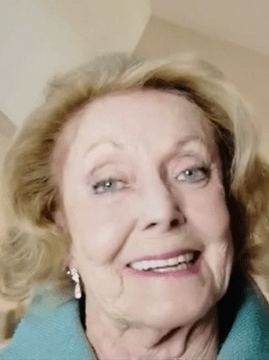 Actress Judith McConnell smiling warmly, wearing a light blue top and earrings, in a softly lit indoor setting.