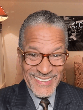 Actor Phil Morris smiling in a pinstriped suit and tie, seated indoors with decorative pillows and a framed photo in the background.